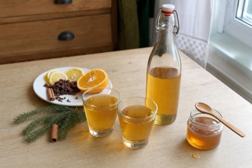 Domáca medovina zlatej farby v dvoch malých pohároch, vedľa fľaša medoviny a pohár medu na drevenom stole